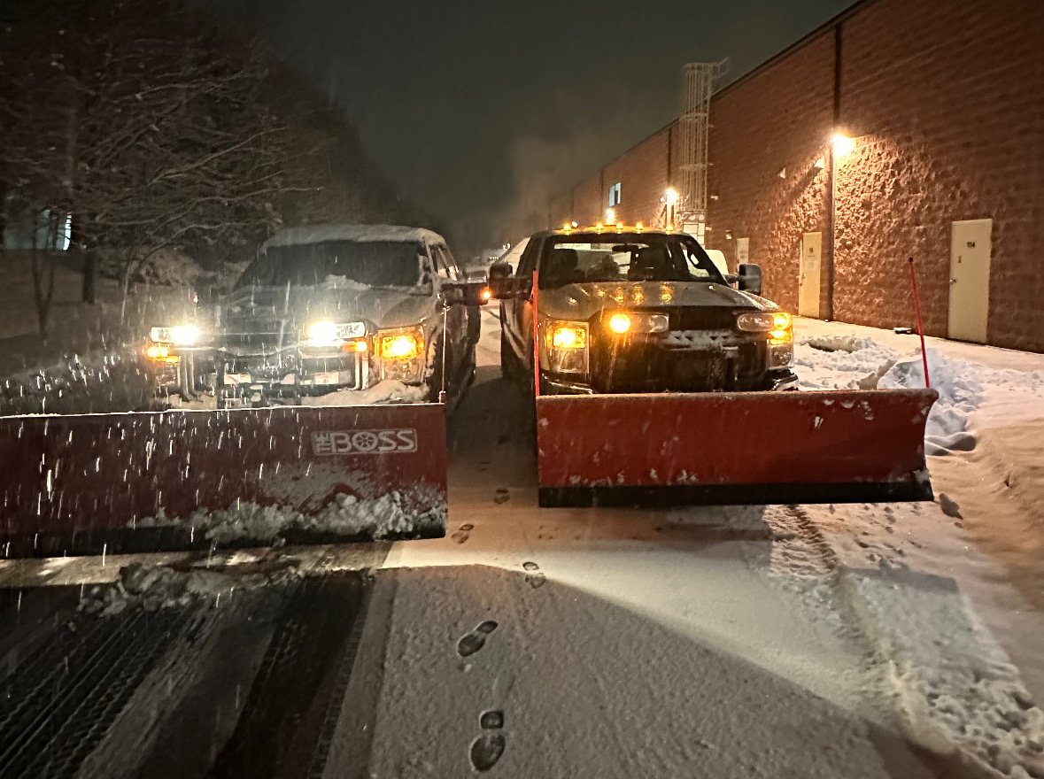 Two plow trucks side by side clearing the service lane behind a commercial building at night